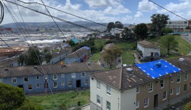 Urban slope: a view from above of residential buildings with tarped roof, overlooking an industrial area, under a cloudy sky with power lines crisscrossing overhead.