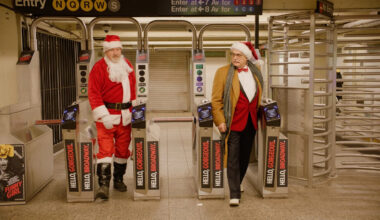 Two men, one dressed as Santa and one in a tan jacket with a Santa hat, stand next to turnstiles in a subway station. A "Funny Girl" poster is visible on the left.