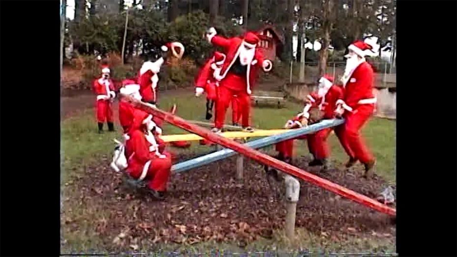 A group of people dressed as Santa Claus play on seesaws in an outdoor park on a cloudy day.