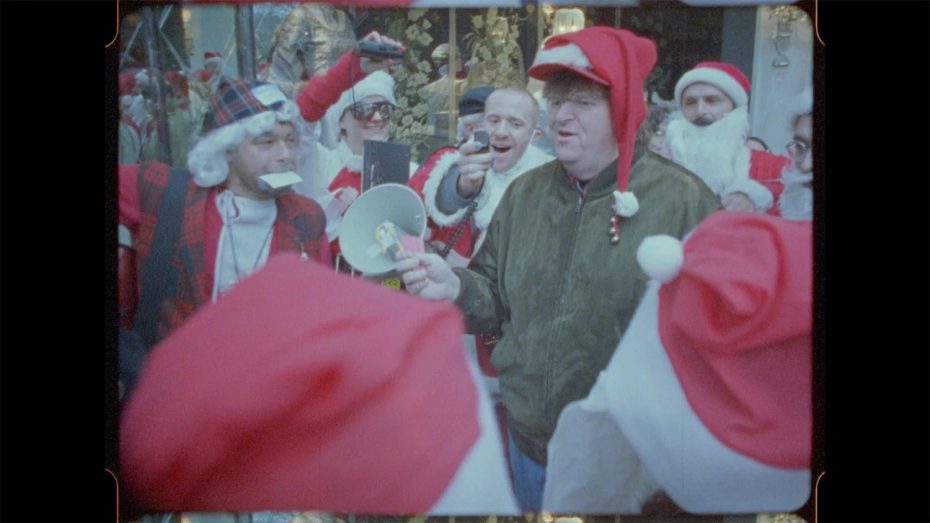 A group of people wearing Santa hats and costumes gather outdoors; one person in the center holds a megaphone and wears a green jacket and red Santa hat.
