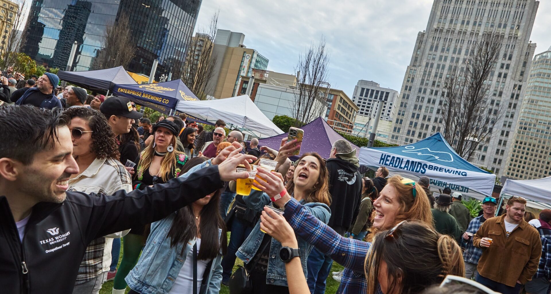 People gathered in a circle, standing outside and raising their glasses as they cheers with beer.