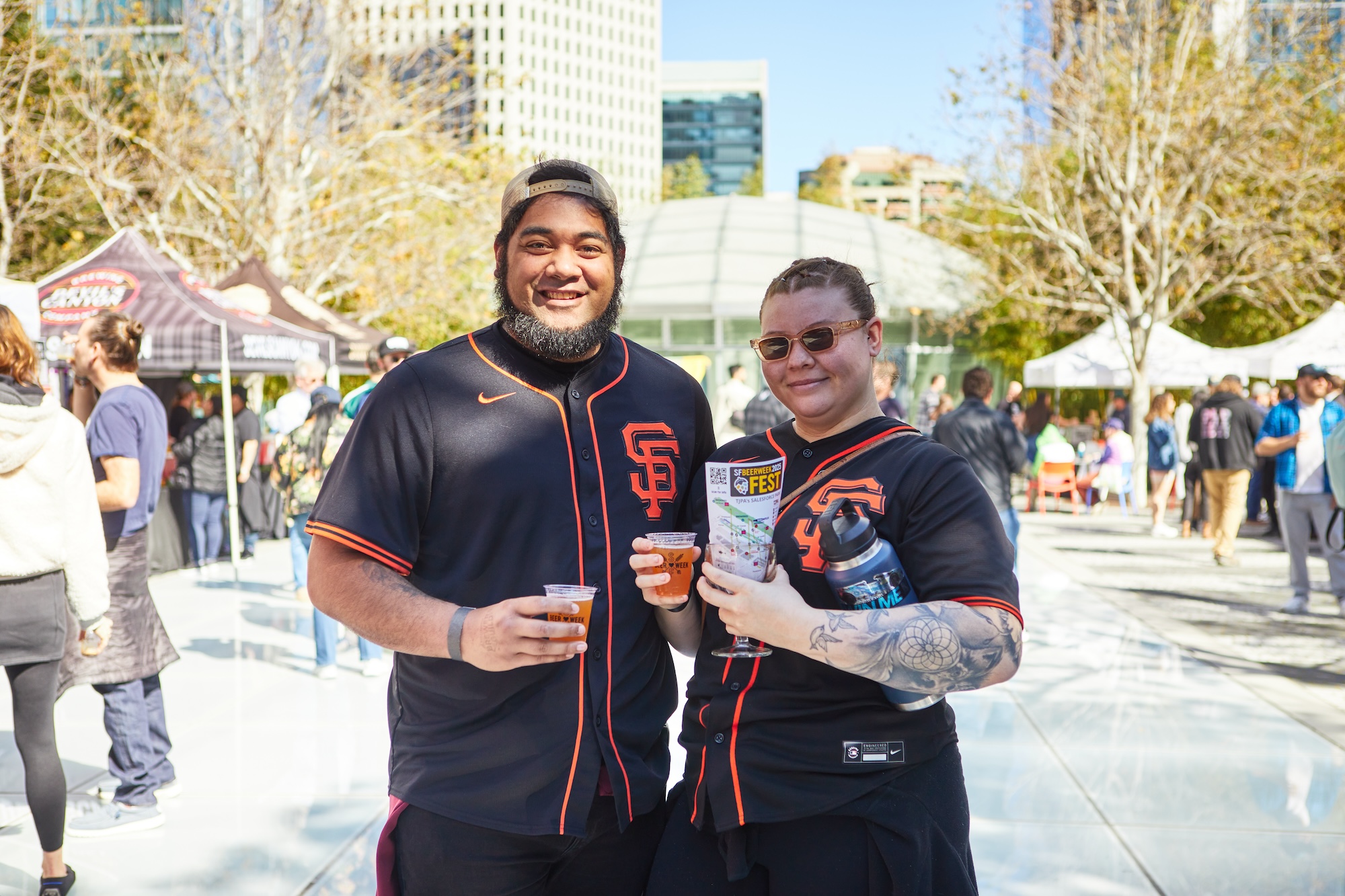 Two people wearing SF Giants gear pose for a photo outside while holding a couple of beers.