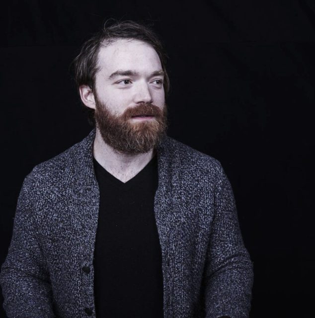 A man with a beard and mustache wearing a gray cardigan over a black shirt stands against a plain dark background.