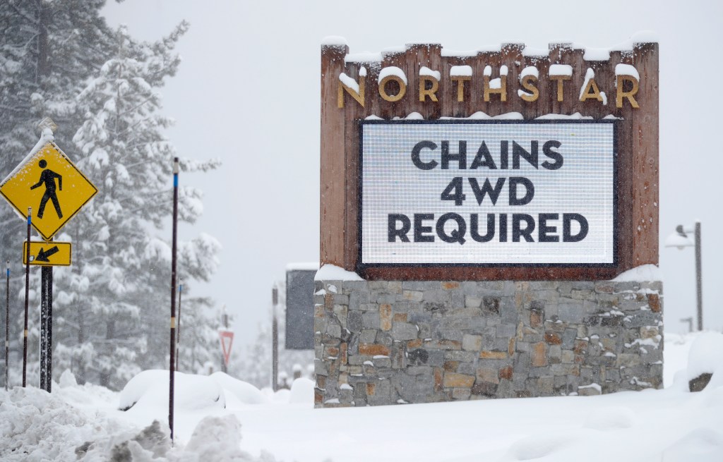 A Northstar California Resort sign covered in snow states "CHAINS 4WD REQUIRED" with a pedestrian crossing sign next to it.