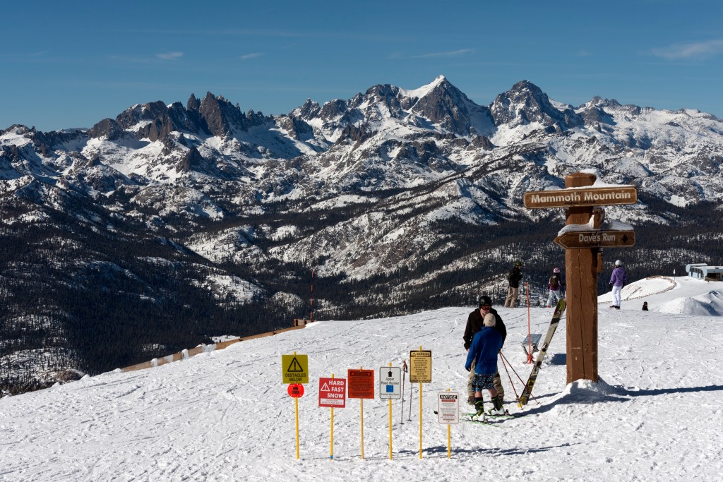 Skiers on Mammoth Mountain, California, with warning signs and mountains in the background.