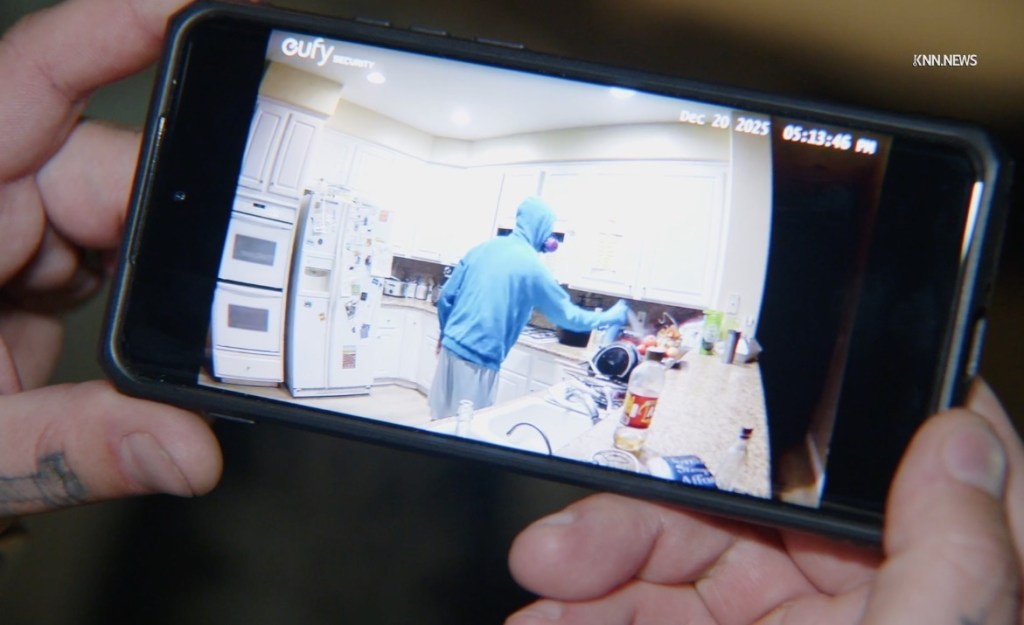 A person in a blue hoodie and respirator mask spraying food in a kitchen, as seen on a phone displaying security camera footage.