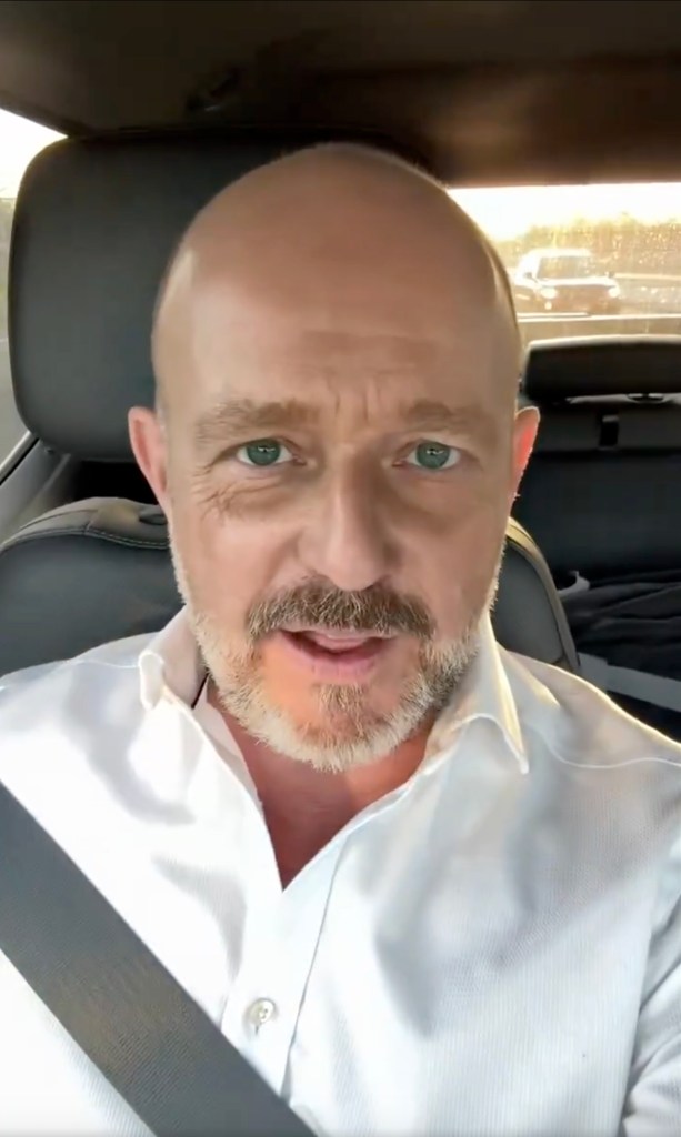 Steve Hilton, wearing a white shirt and seatbelt, looks directly at the camera while speaking in a car.