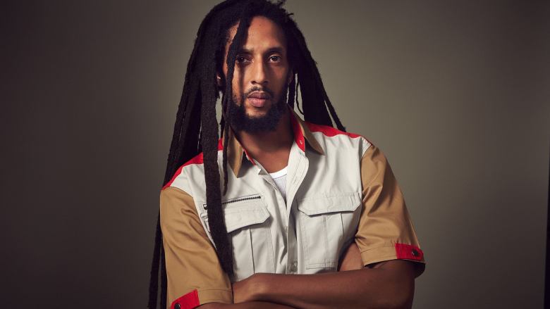 A man with dreadlocks stands with arms folded against a gray background