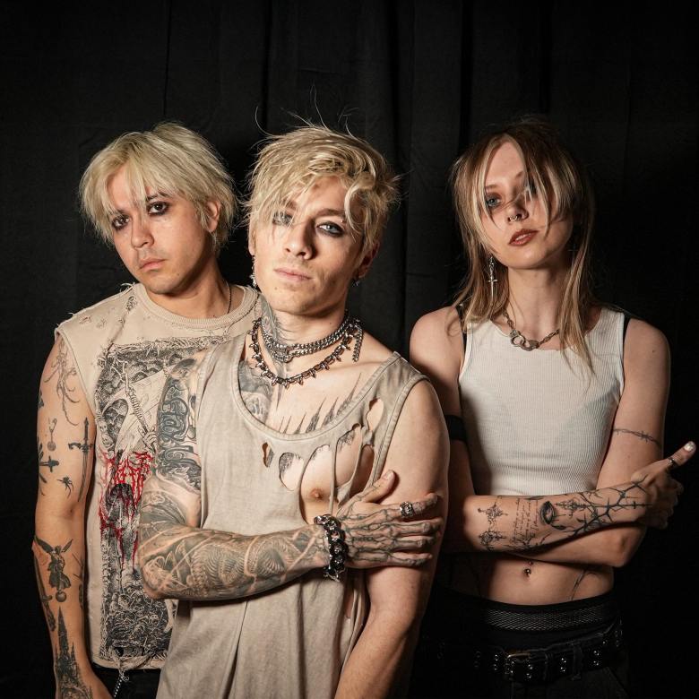 Two men and a girl in with tattoos and wearing tattered clothes stand against a black background