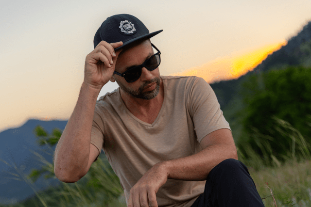 A man poses in sunglasses and a ball cap in a field at sunset