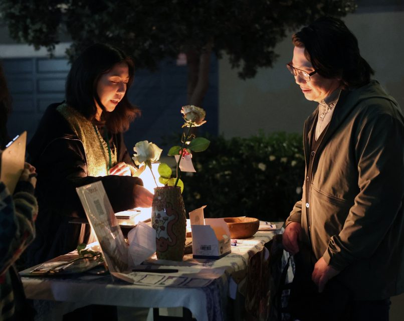 Two people stand at a small outdoor table with papers, flowers, and decorative items, lit by a lamp, at night.