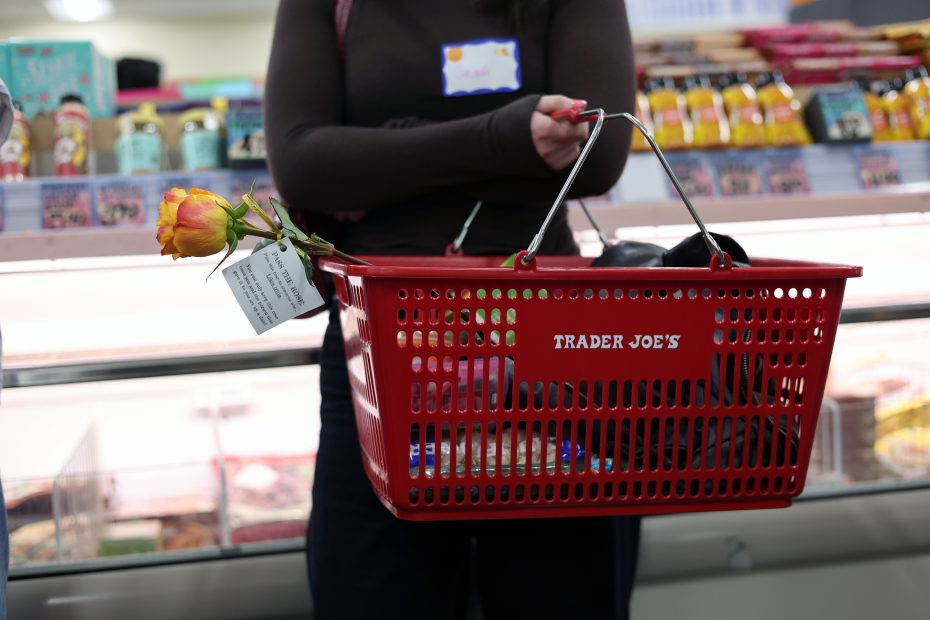 A person holds a red Trader Joe's shopping basket with a single yellow rose and a note inside, standing near a refrigerated display in a grocery store.