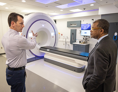 Man with striped shirt motions with hand toward man in dark business suit as they stand in front of a large, cylindrical machine.