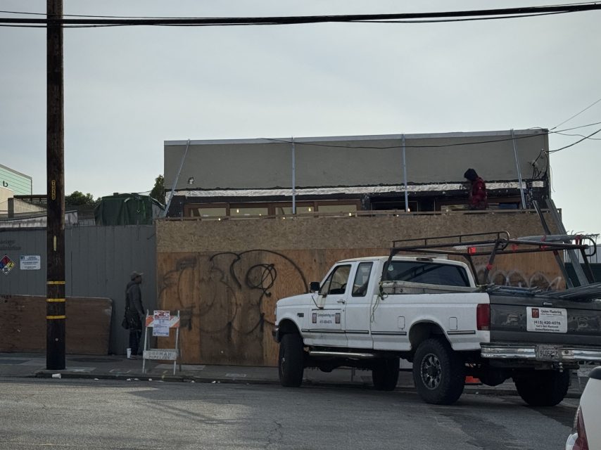 A white pickup truck is parked on the street in front of a construction site with plywood barriers and two people on the partially built structure.
