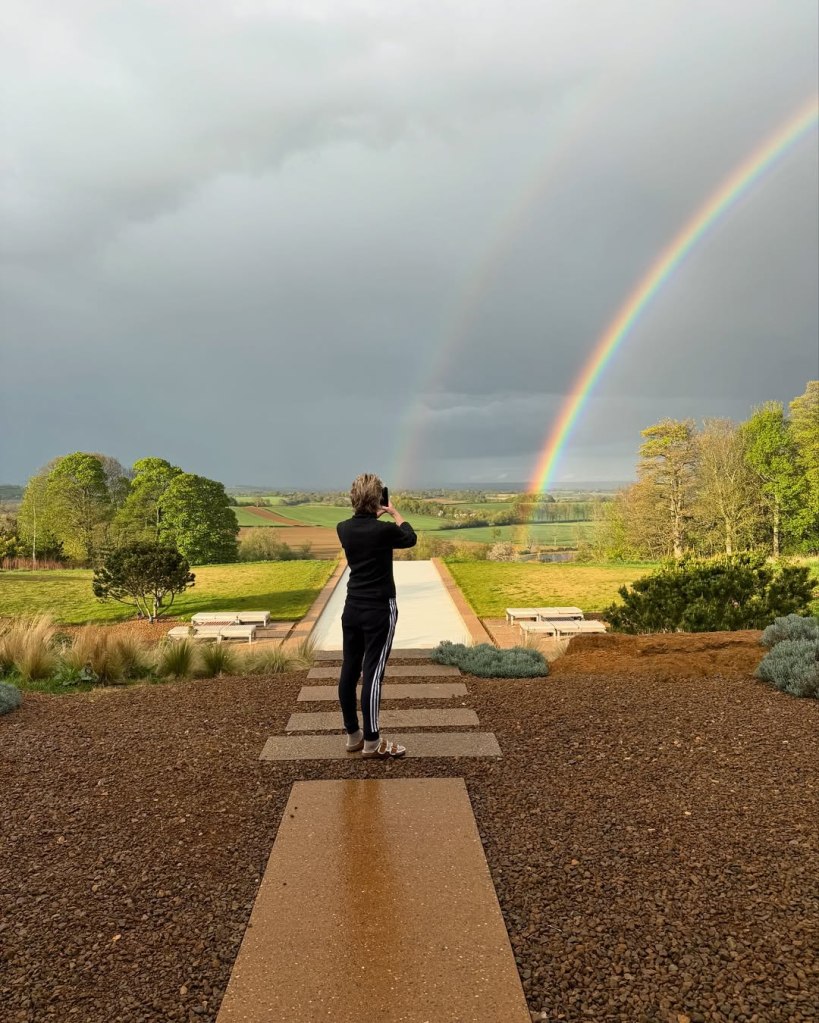 Ellen DeGeneres stands on a stone path, photographing a double rainbow stretching across a cloudy sky over a green landscape.