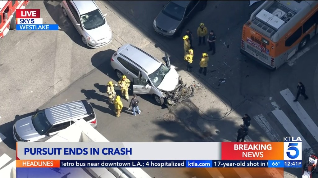 Overhead view of a silver SUV with significant front-end damage after crashing, surrounded by firefighters, police officers, and a Metro bus.