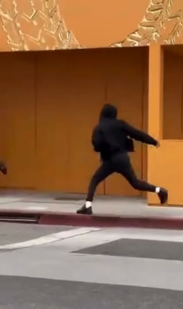 A person dressed in all black, including a hoodie, runs across a street in front of a building with an orange-yellow facade.