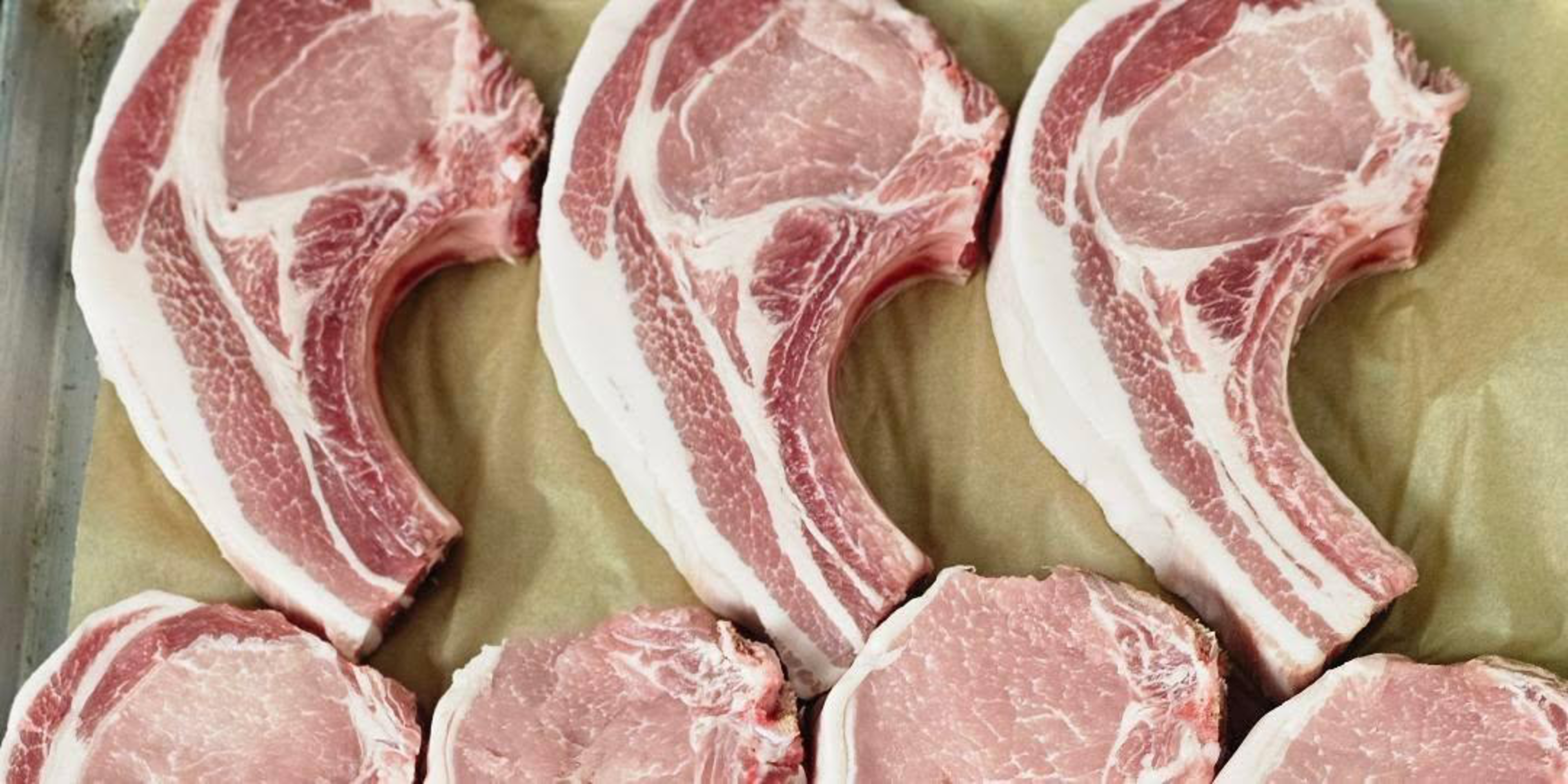 Six raw pork chops with bones are arranged on a sheet of parchment paper, displaying pink meat and white fat marbling.