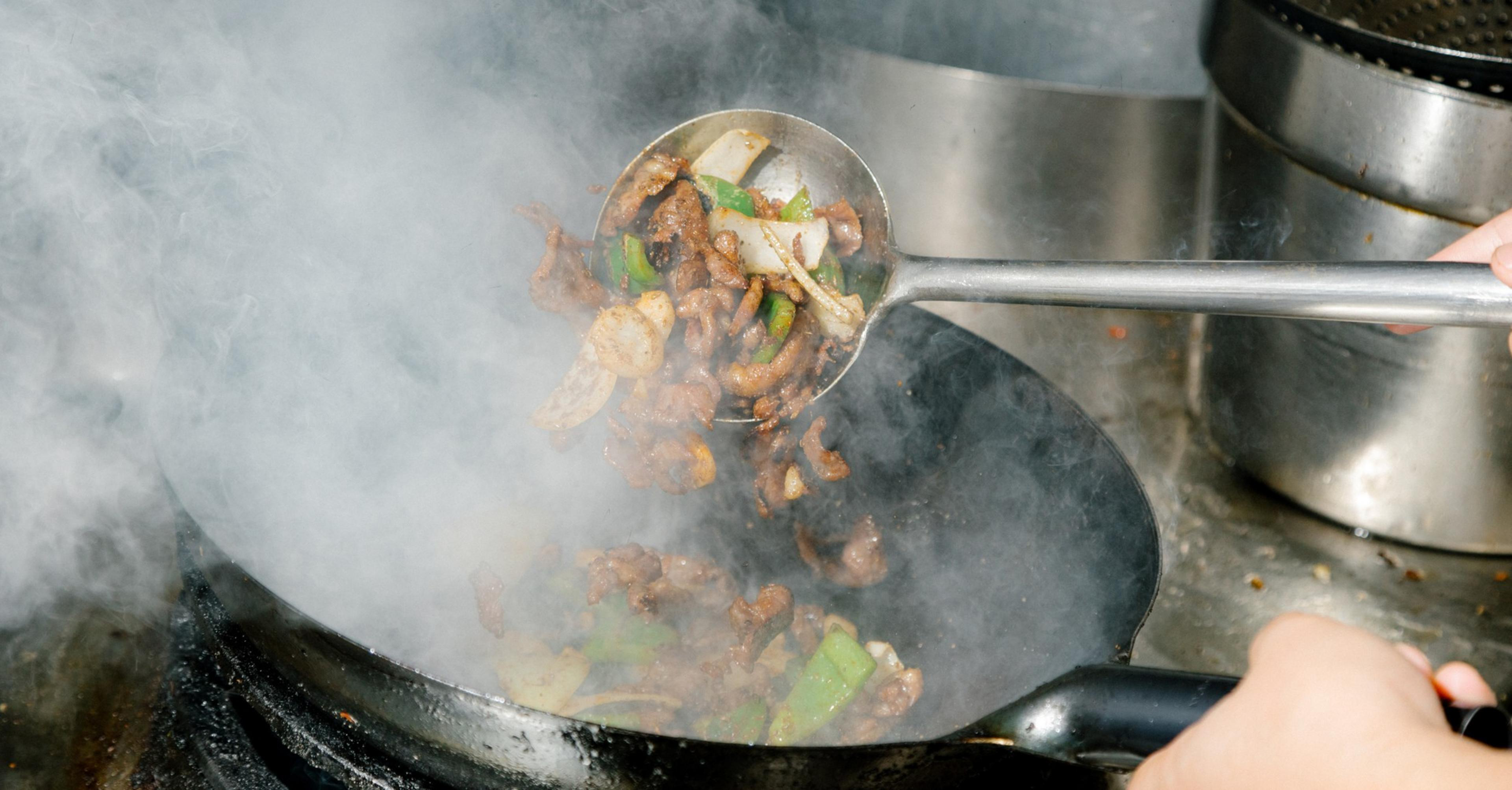 Sizzling stir-fry with meat, onions, and green peppers is being cooked in a smoky wok, with a hand holding a metal ladle to toss the ingredients.