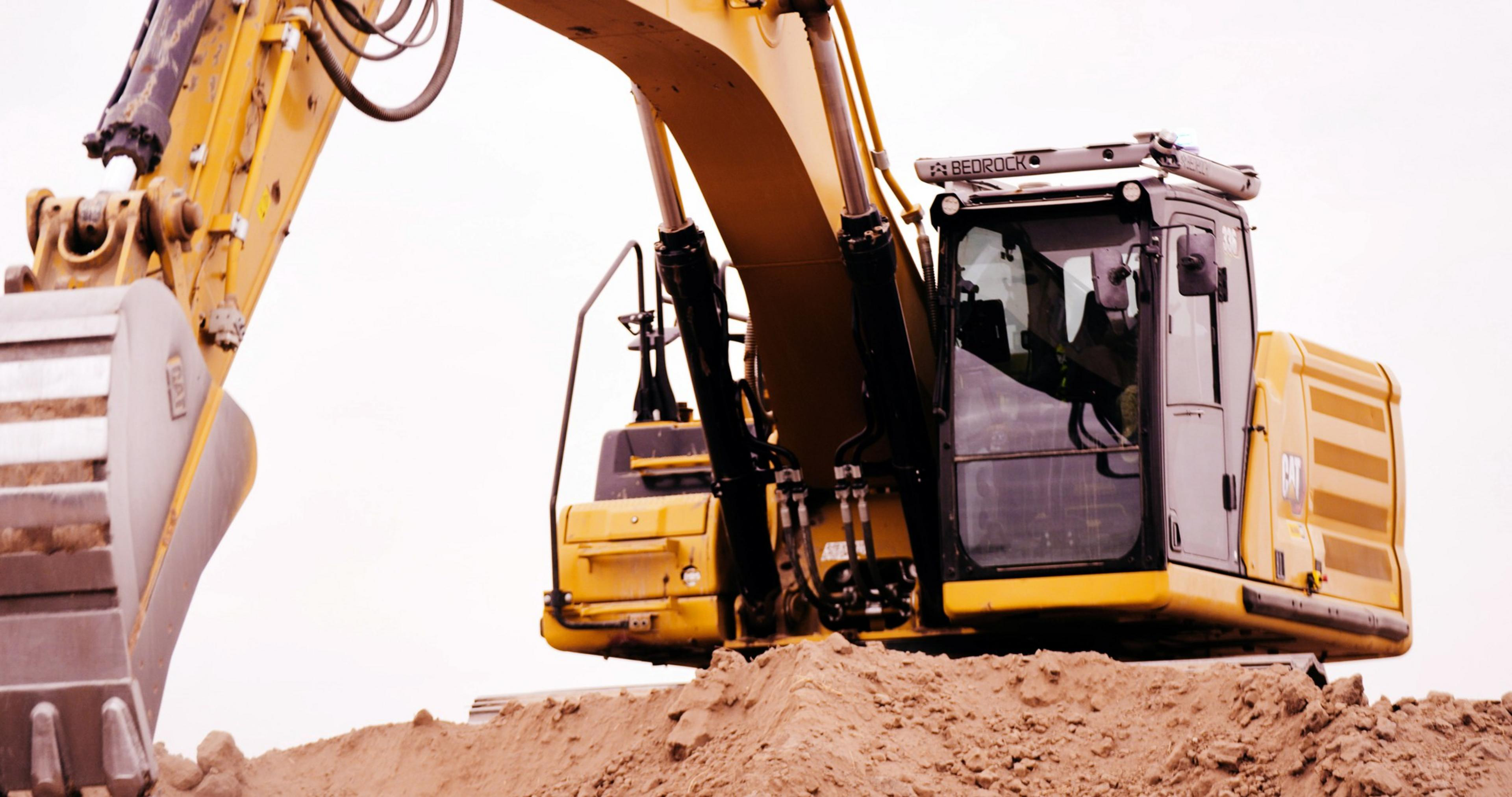 A yellow excavator with a large metal bucket is positioned on top of a mound of dirt against a pale sky.