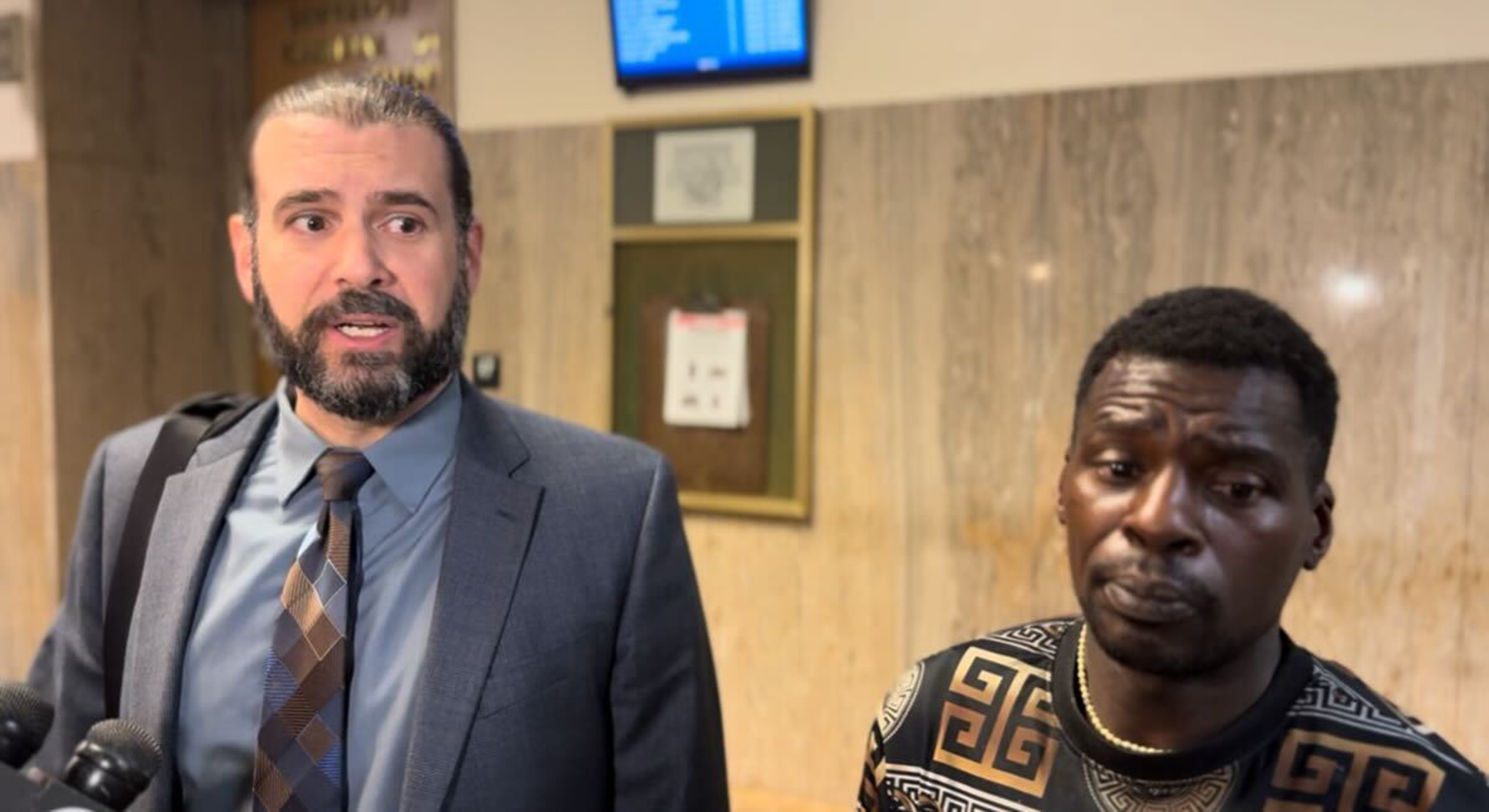 A man in a suit with a beard and tied-back hair speaks, while another man in a patterned shirt and necklace listens quietly beside him.
