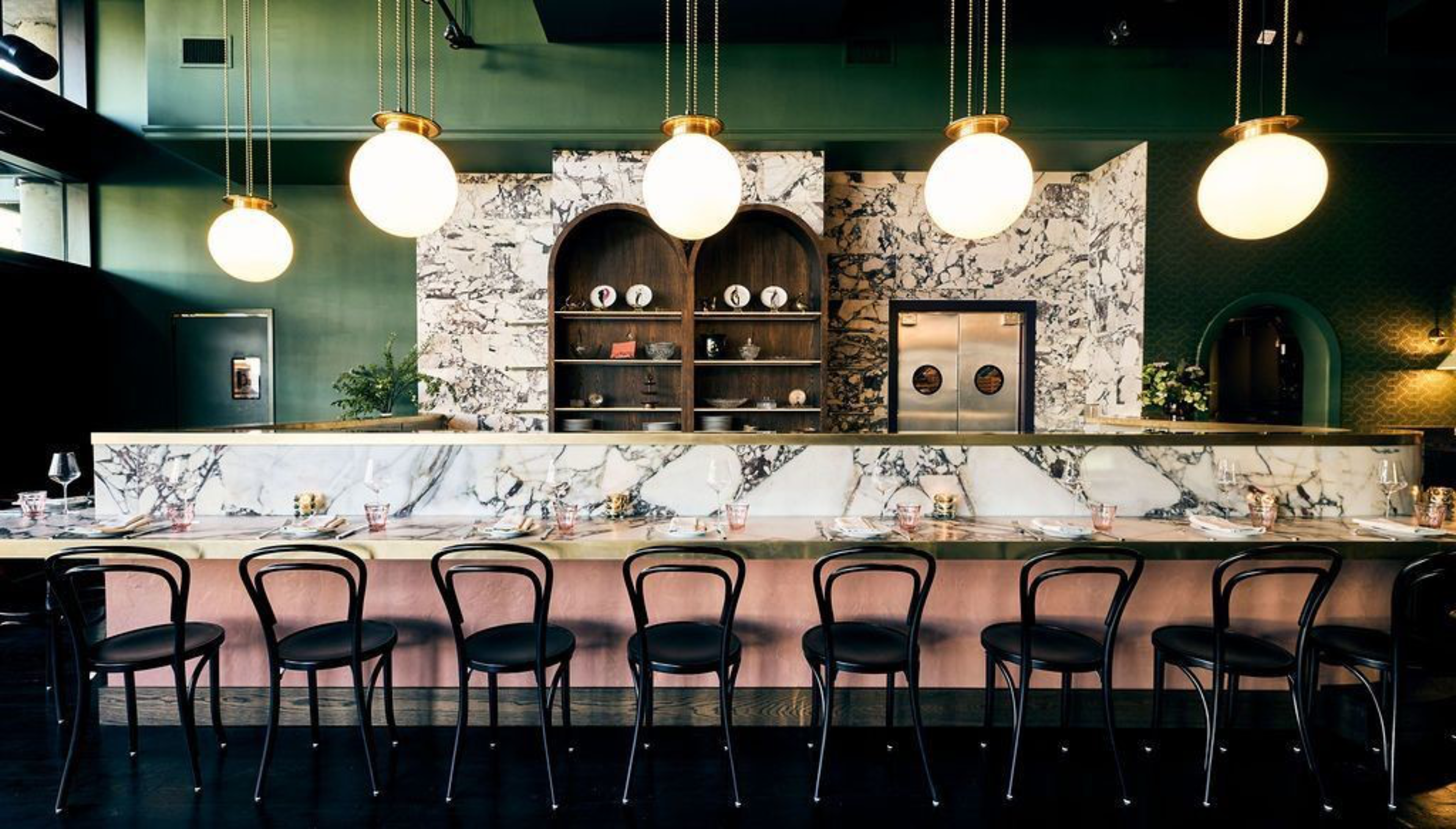 A sleek bar with a marble countertop, eight black chairs, round hanging lights, and a marble wall with wooden shelves holding plates behind.