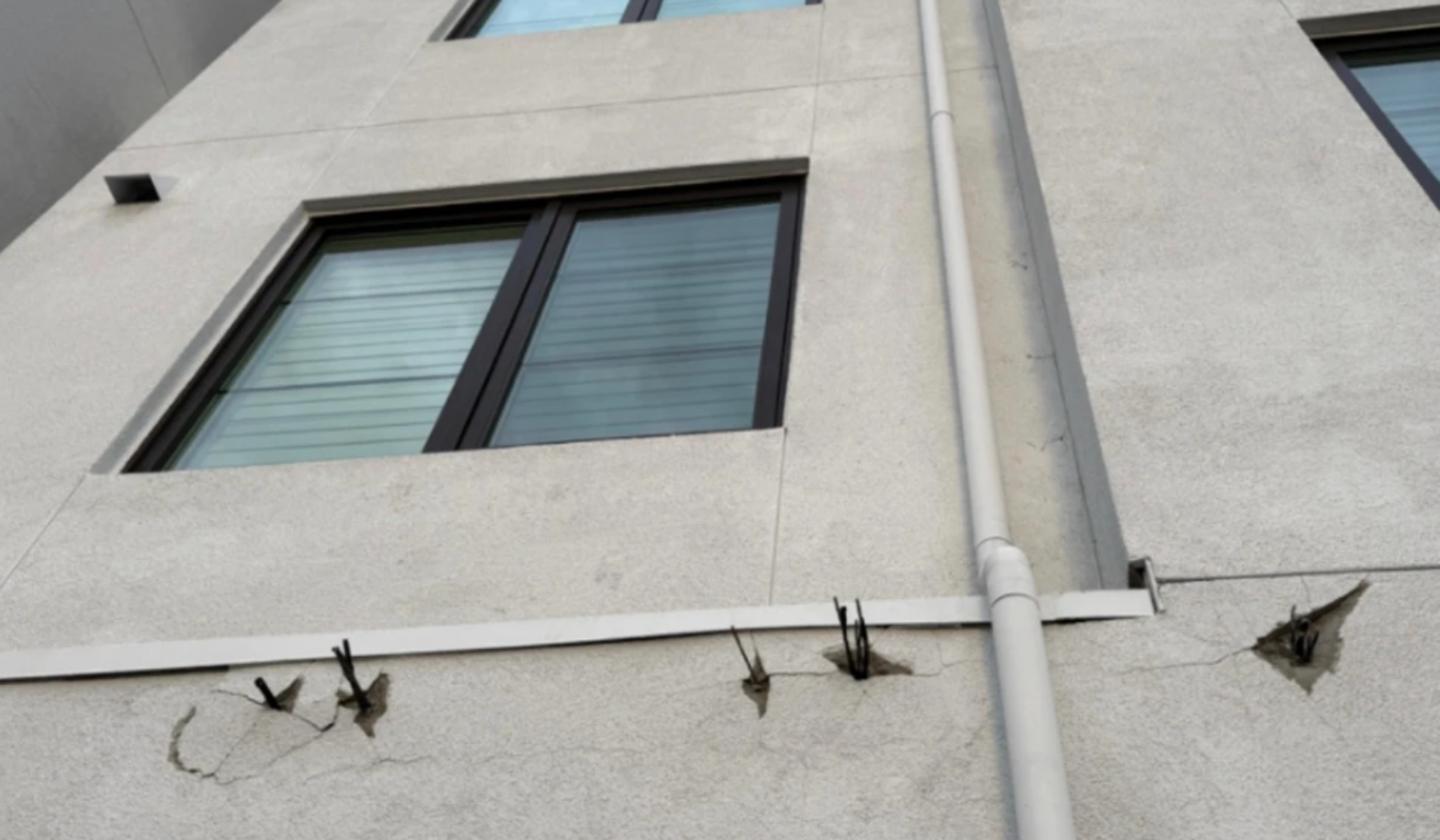 A beige building wall with two windows and several exposed pieces of rebar protruding from cracked concrete near a vertical white pipe.