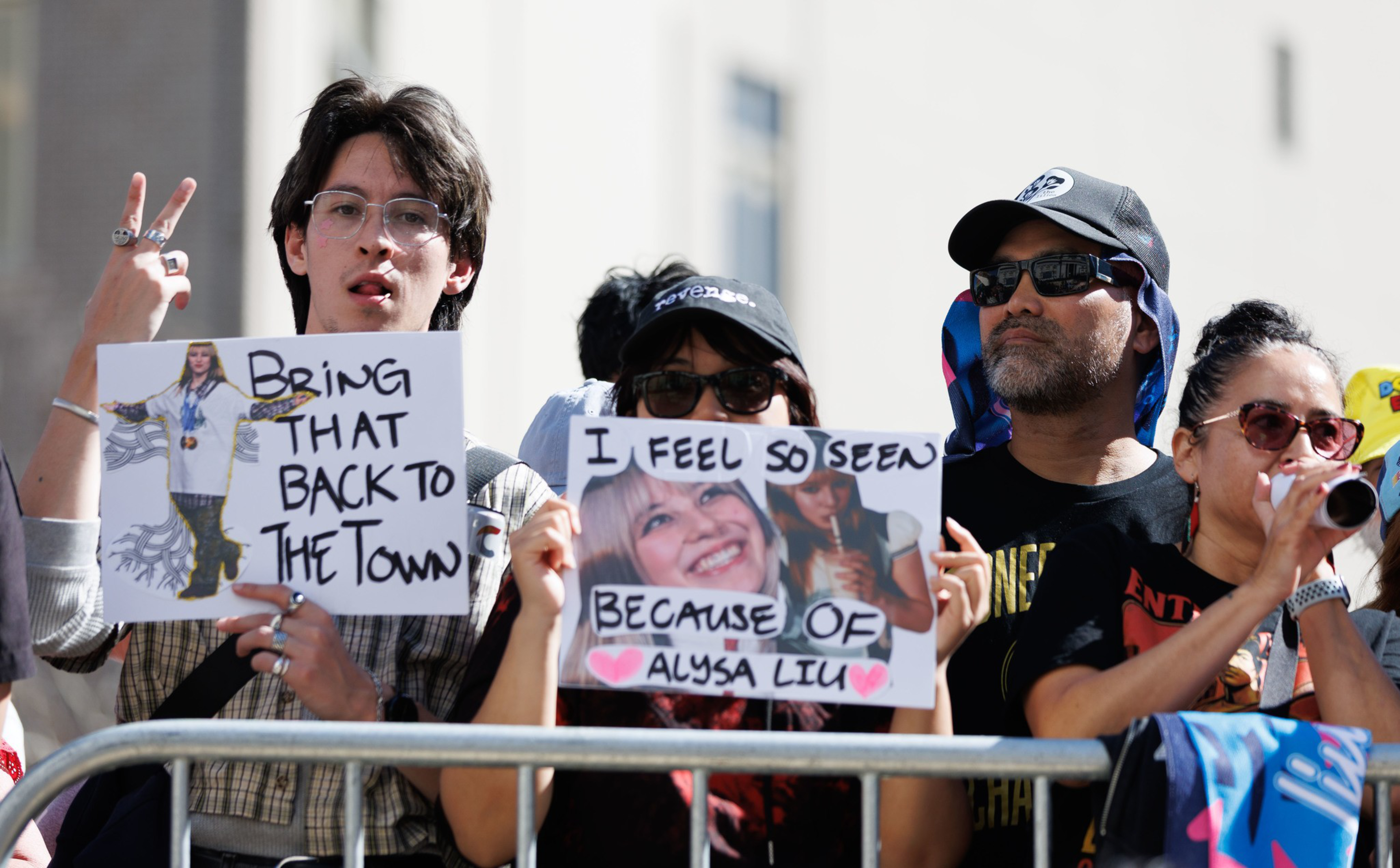 Four people stand behind a metal barrier holding signs, one reading “BRING THAT BACK TO THE TOWN” and another “I FEEL SO SEEN BECAUSE OF ALYSA LIU.”