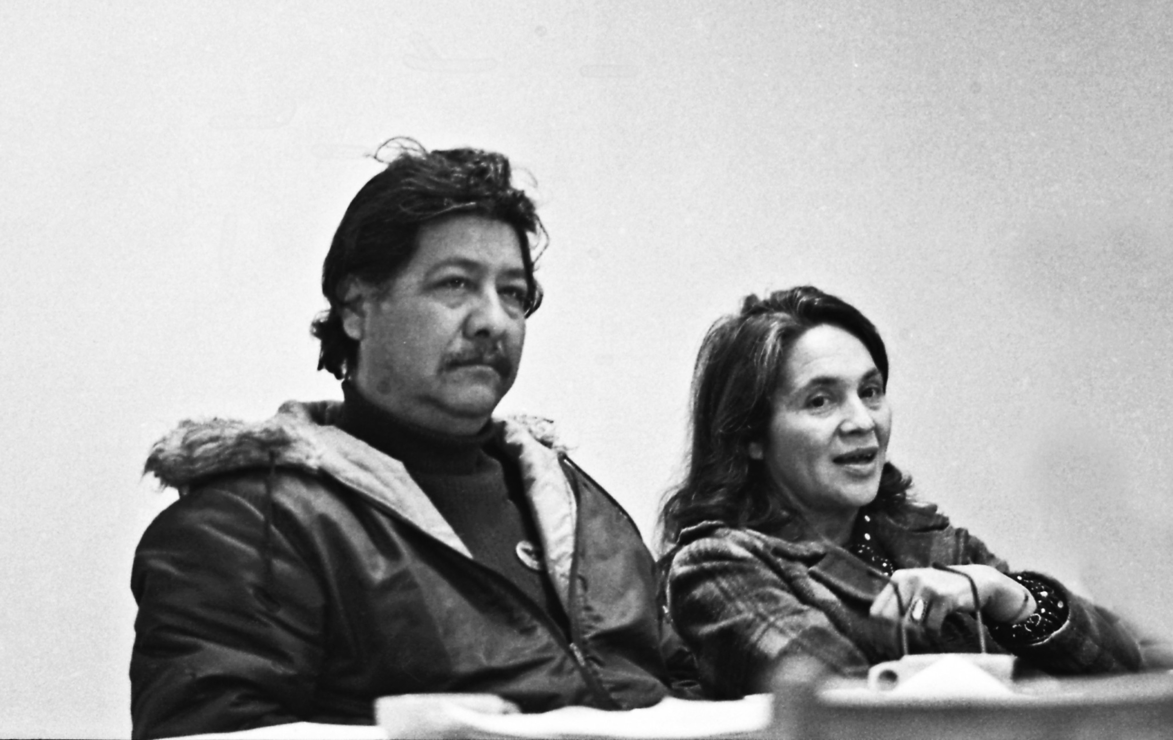 A man in a heavy jacket with a fur-lined hood sits next to a woman wearing a plaid coat, both appearing to be attentively listening or speaking.