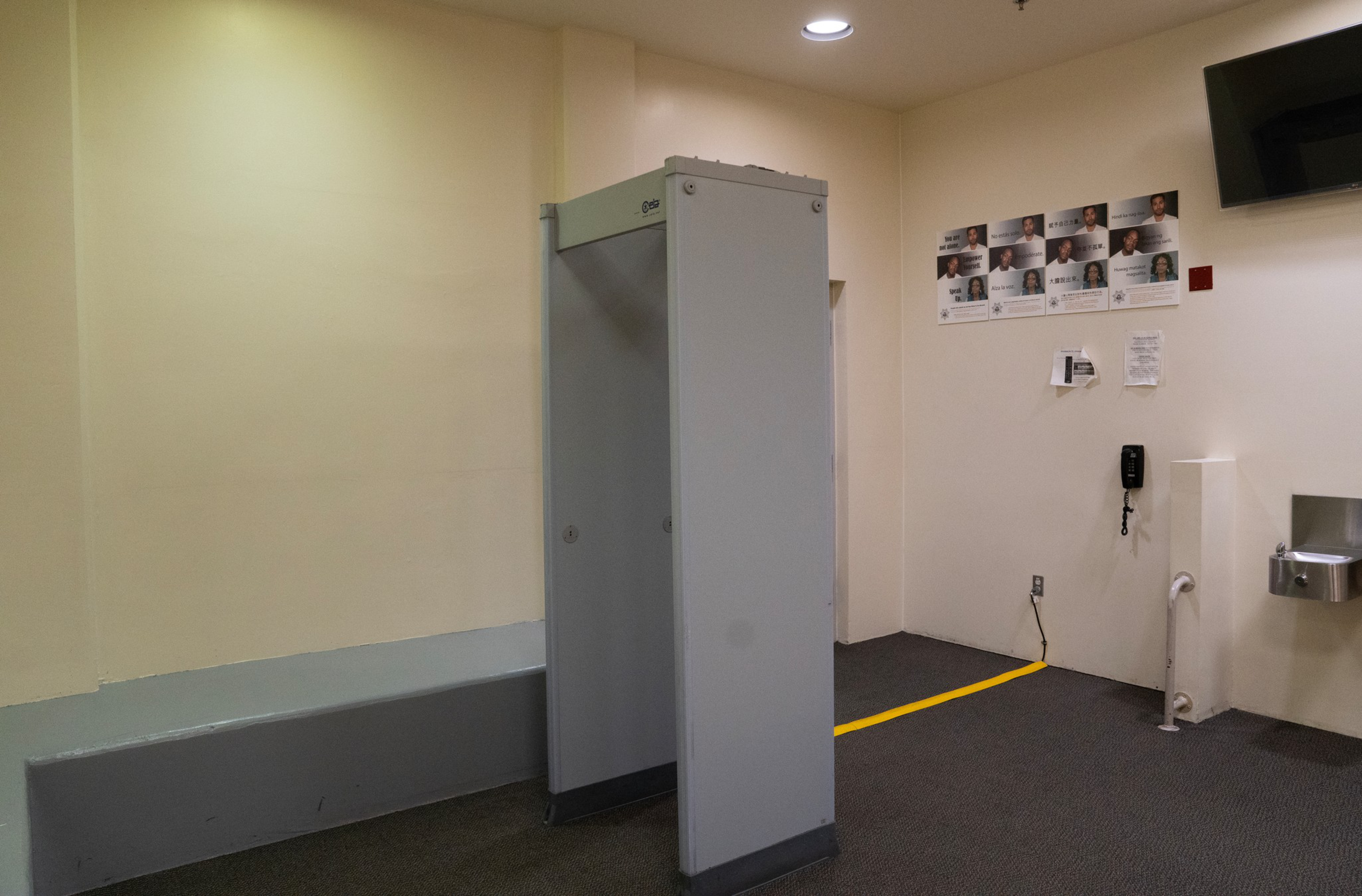 A gray metal detector stands near a beige wall with a bench, a water fountain, a telephone, and a wall displaying photos of people.