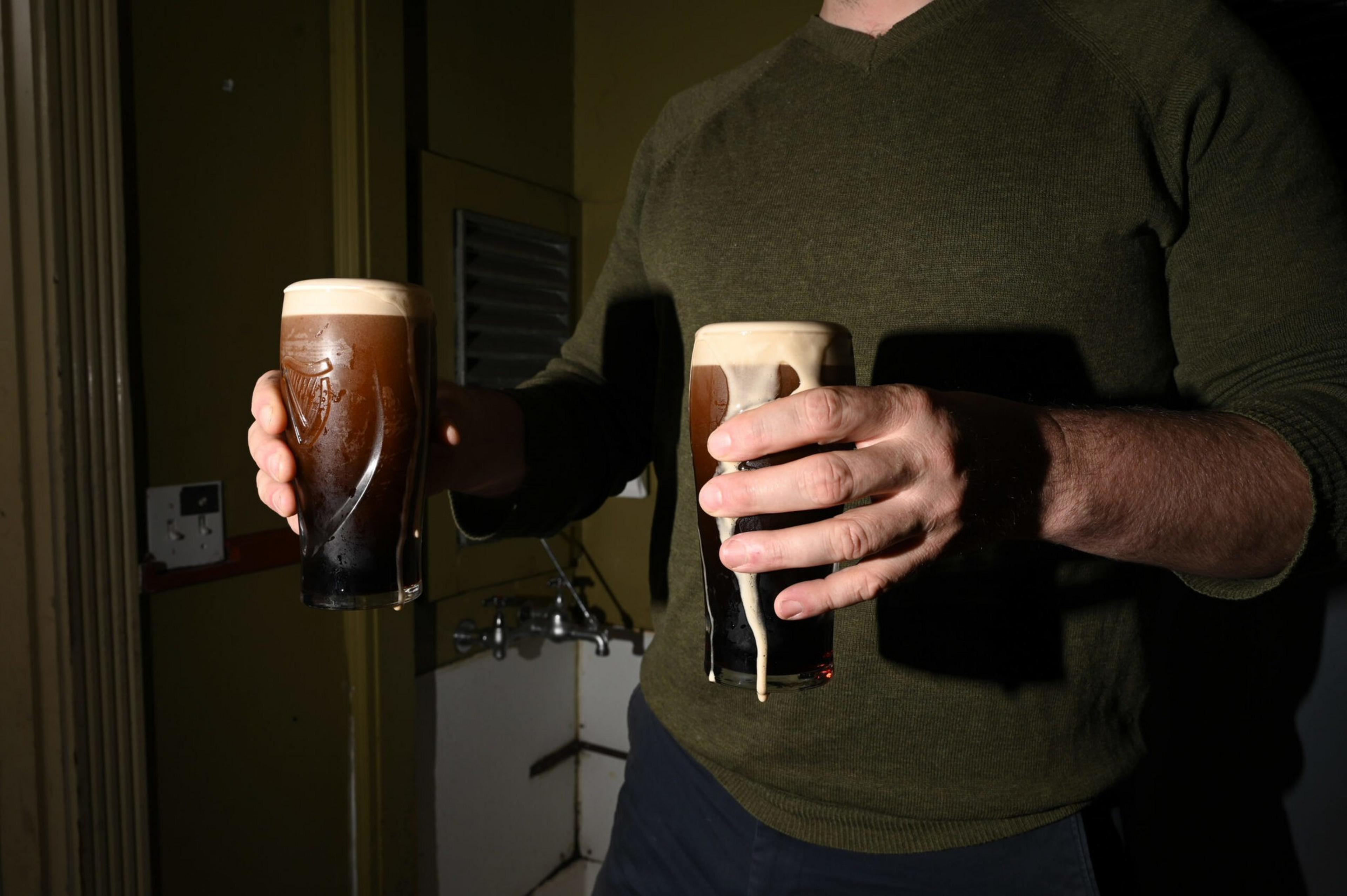 A person wearing a dark green sweater holds two glasses of dark beer with creamy foam, one glass having some foam dripping down the side.