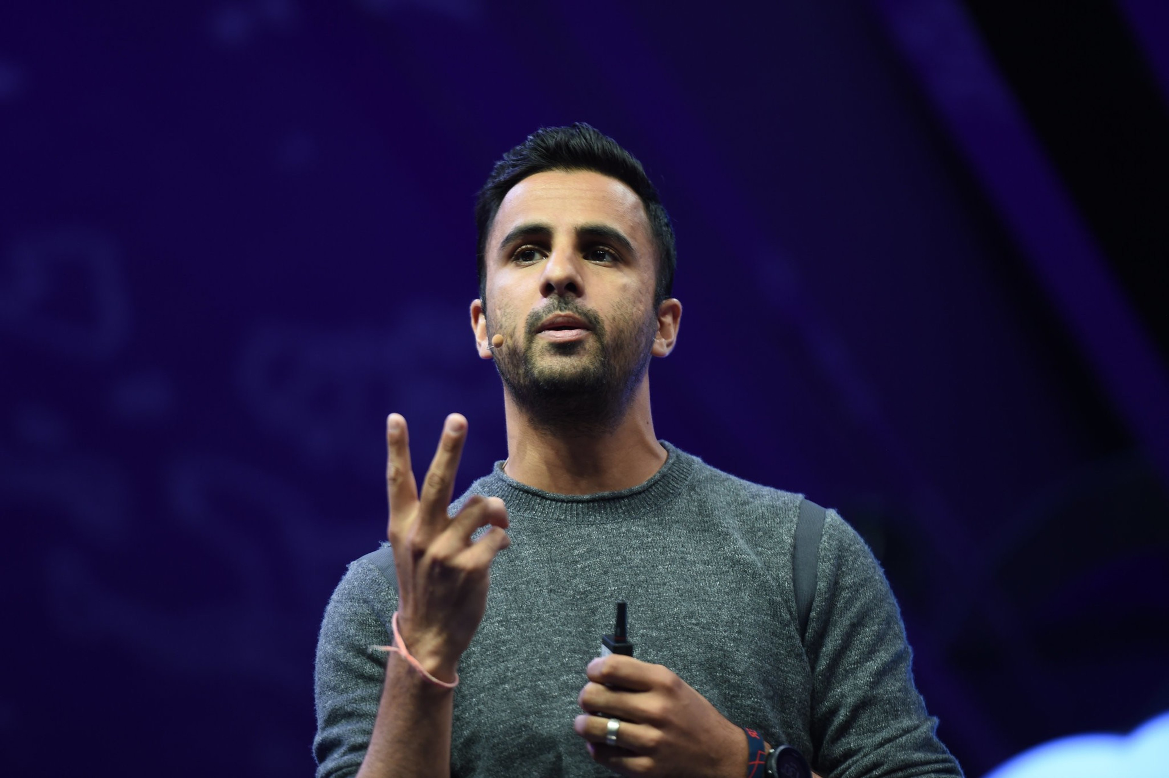 A man with short dark hair and a beard speaks, wearing a gray sweater and holding a microphone, with a dark purple background behind him.