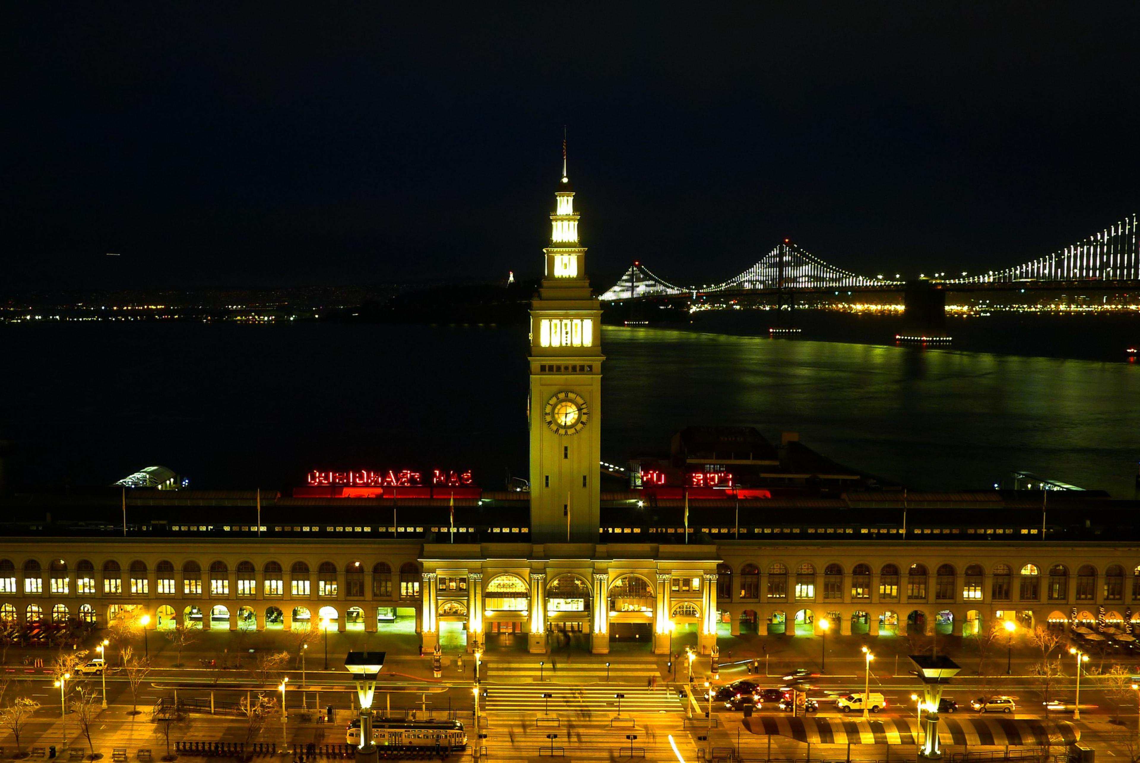 A brightly lit clock tower stands at the center of a large building by the water, with a lit suspension bridge and city lights in the background at night.