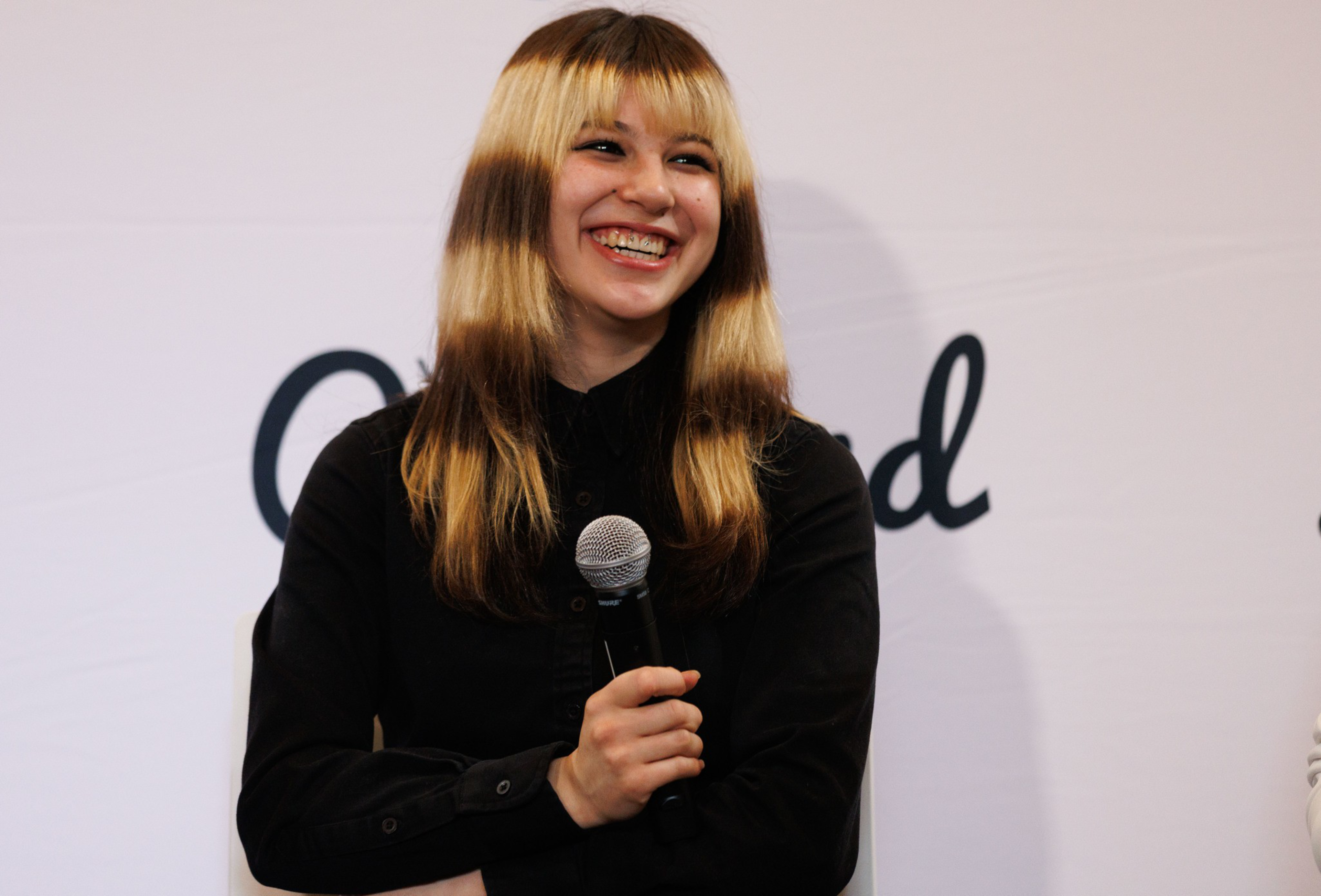 A smiling young woman with blonde hair and dark roots holds a microphone, wearing a black shirt against a light background with partial text.