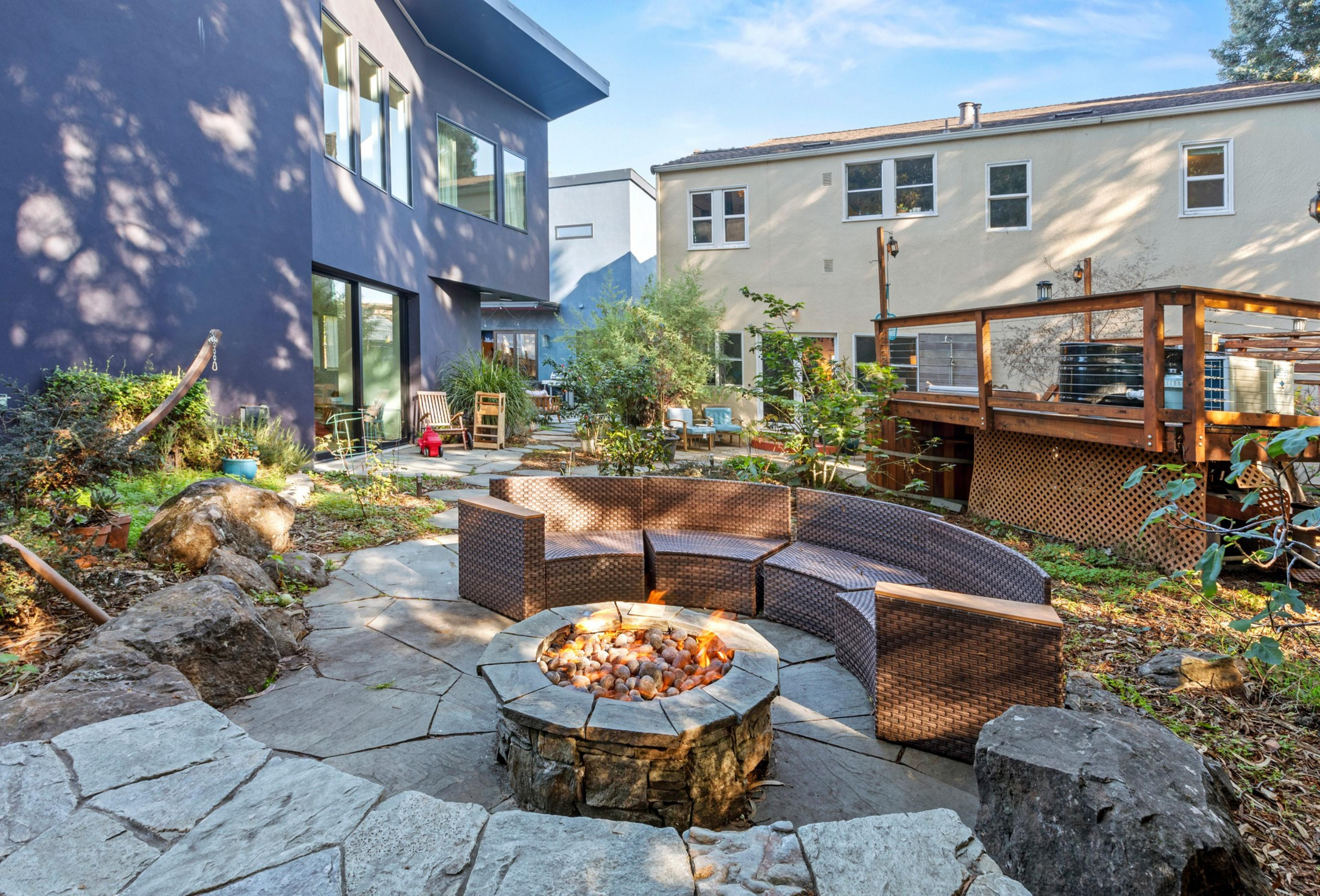Curved wicker seating surrounds a stone fire pit on a patio, with modern blue and beige houses and a wooden deck in the background under a clear sky.