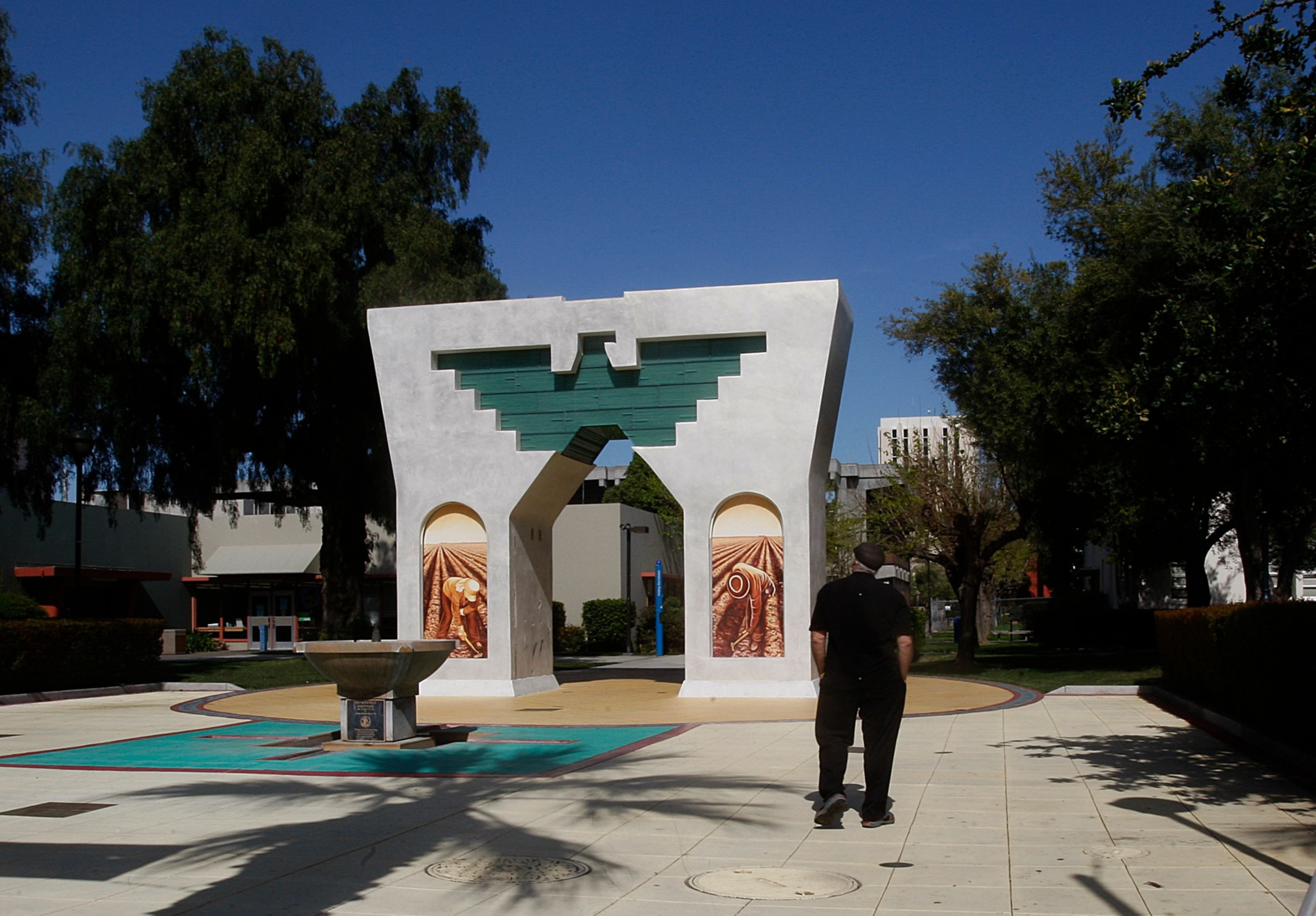 A man walks toward a white, arch-shaped monument with green and brown geometric designs, surrounded by trees and a clear blue sky.