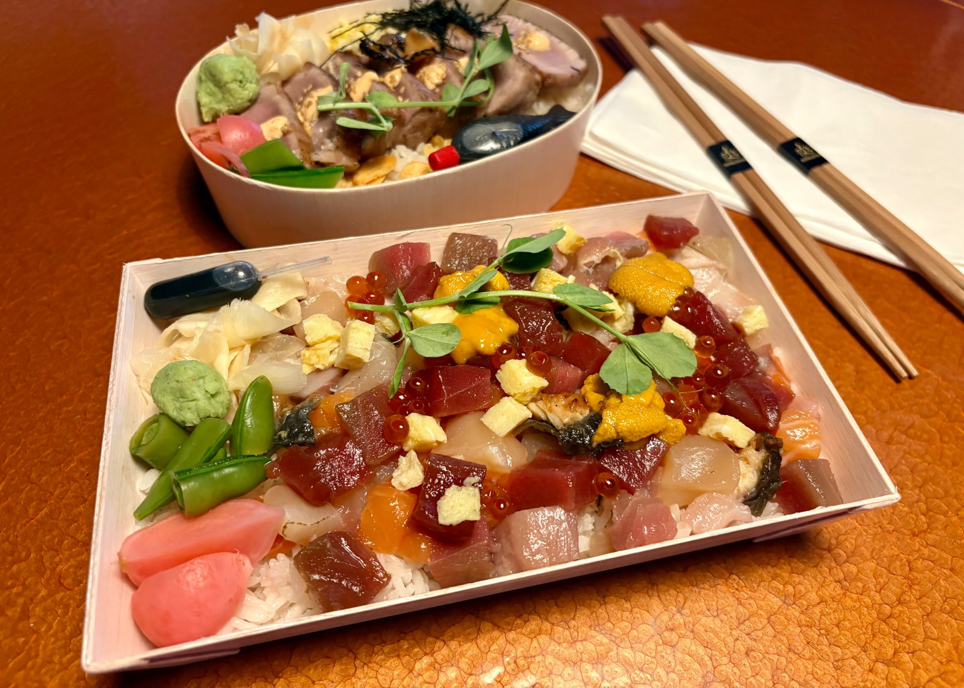 Two bento boxes with assorted sashimi, rice, garnishes like wasabi, pickled ginger, and peas, with chopsticks and napkin on the side.