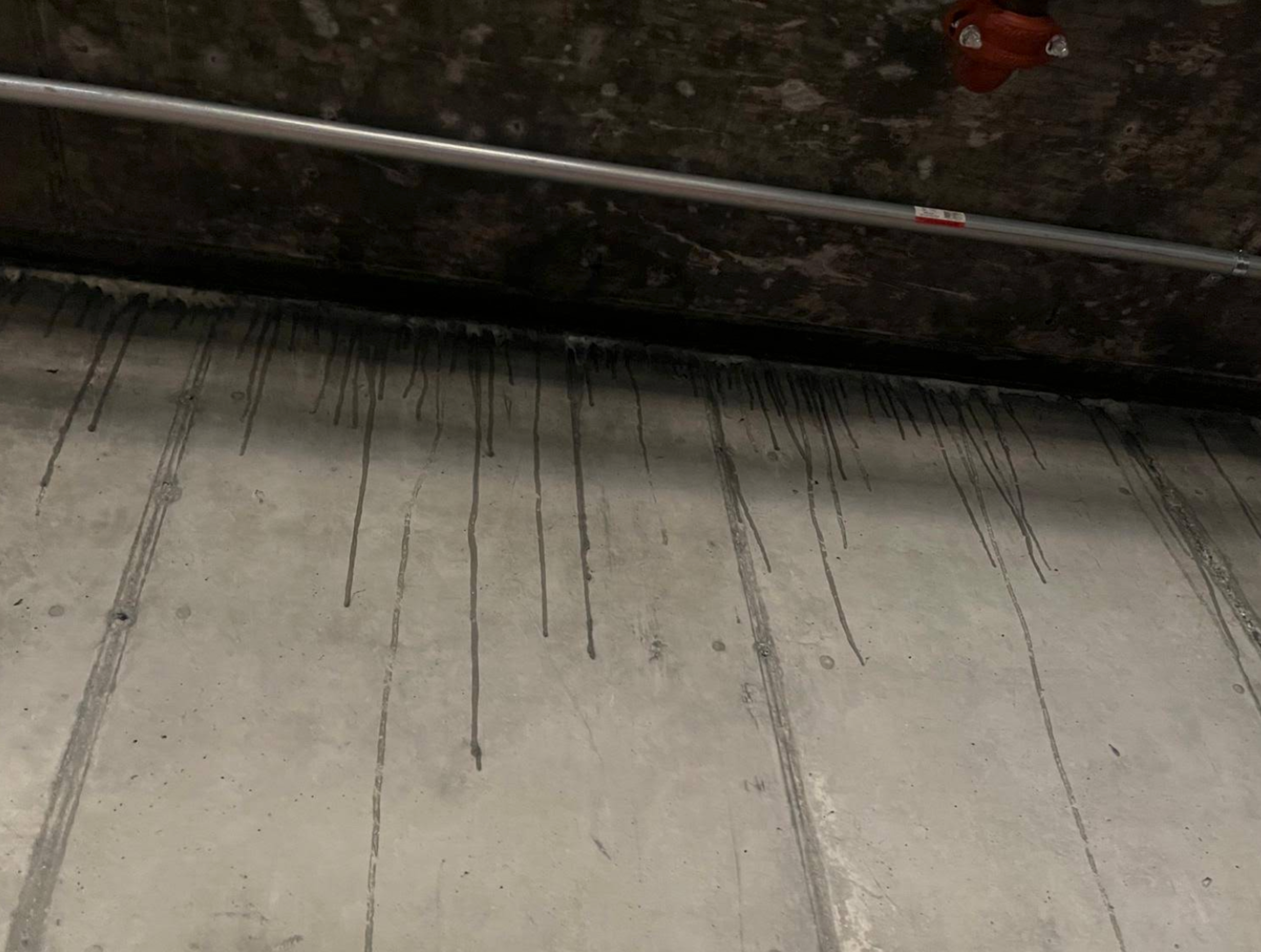 Dark streaks run down a concrete wall from where it meets a darker ceiling with an attached metal pipe and red valve.
