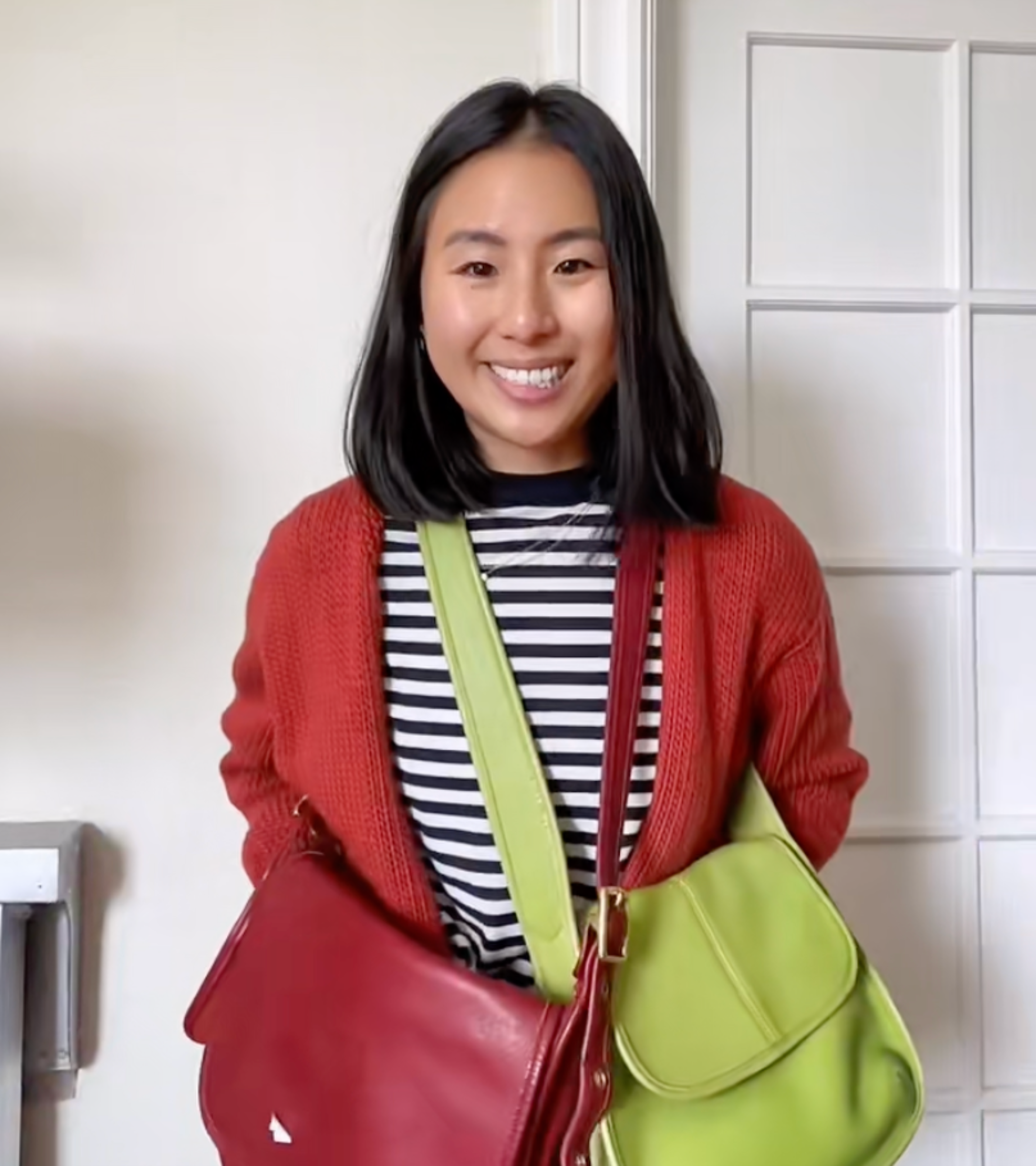 A smiling woman with black hair wears a red cardigan over a black-and-white striped shirt, holding a red bag and a green bag crossed over her chest.