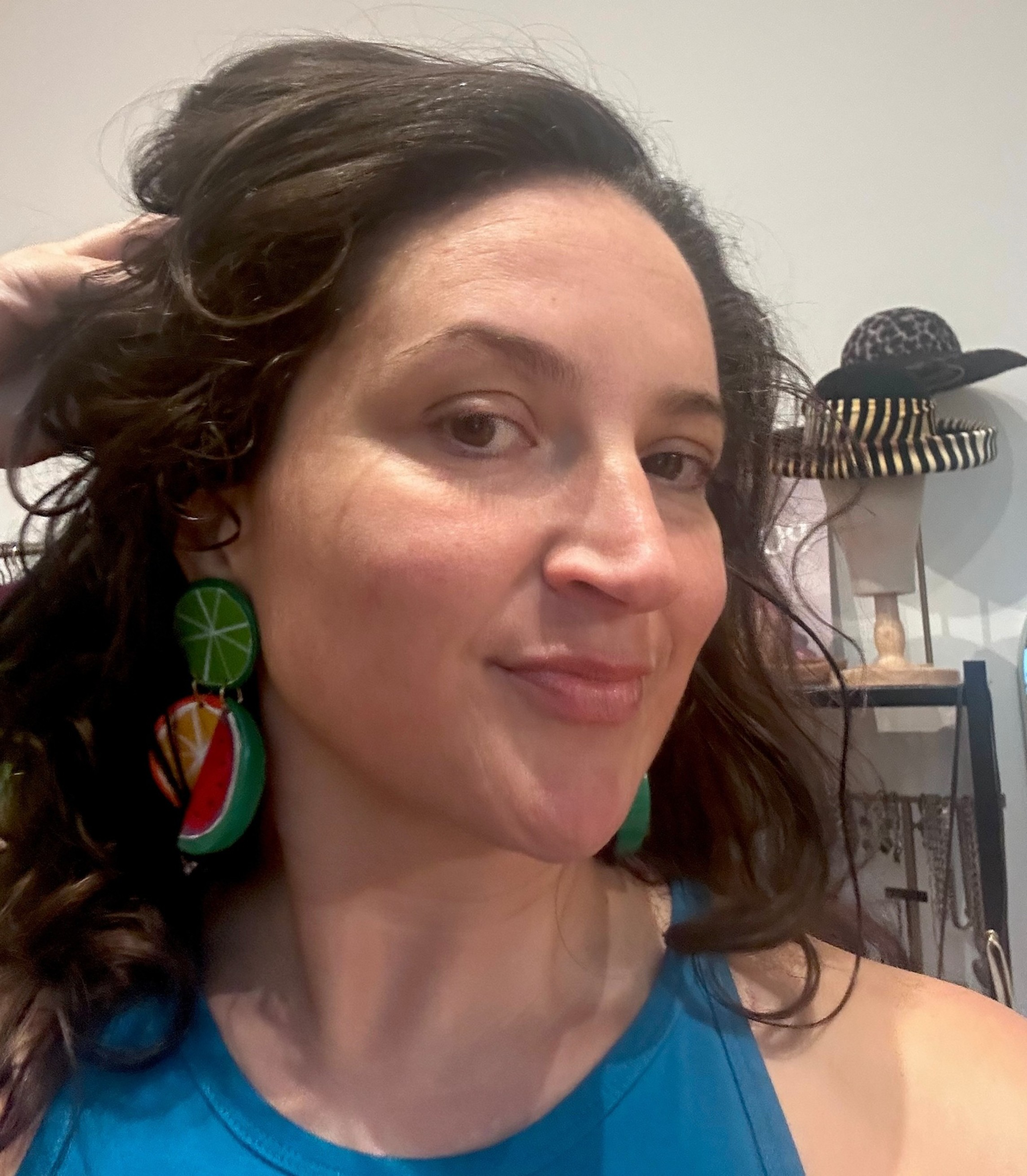 A woman with wavy brown hair wears colorful fruit-shaped earrings and a blue top, smiling slightly with shelves and hats visible in the background.