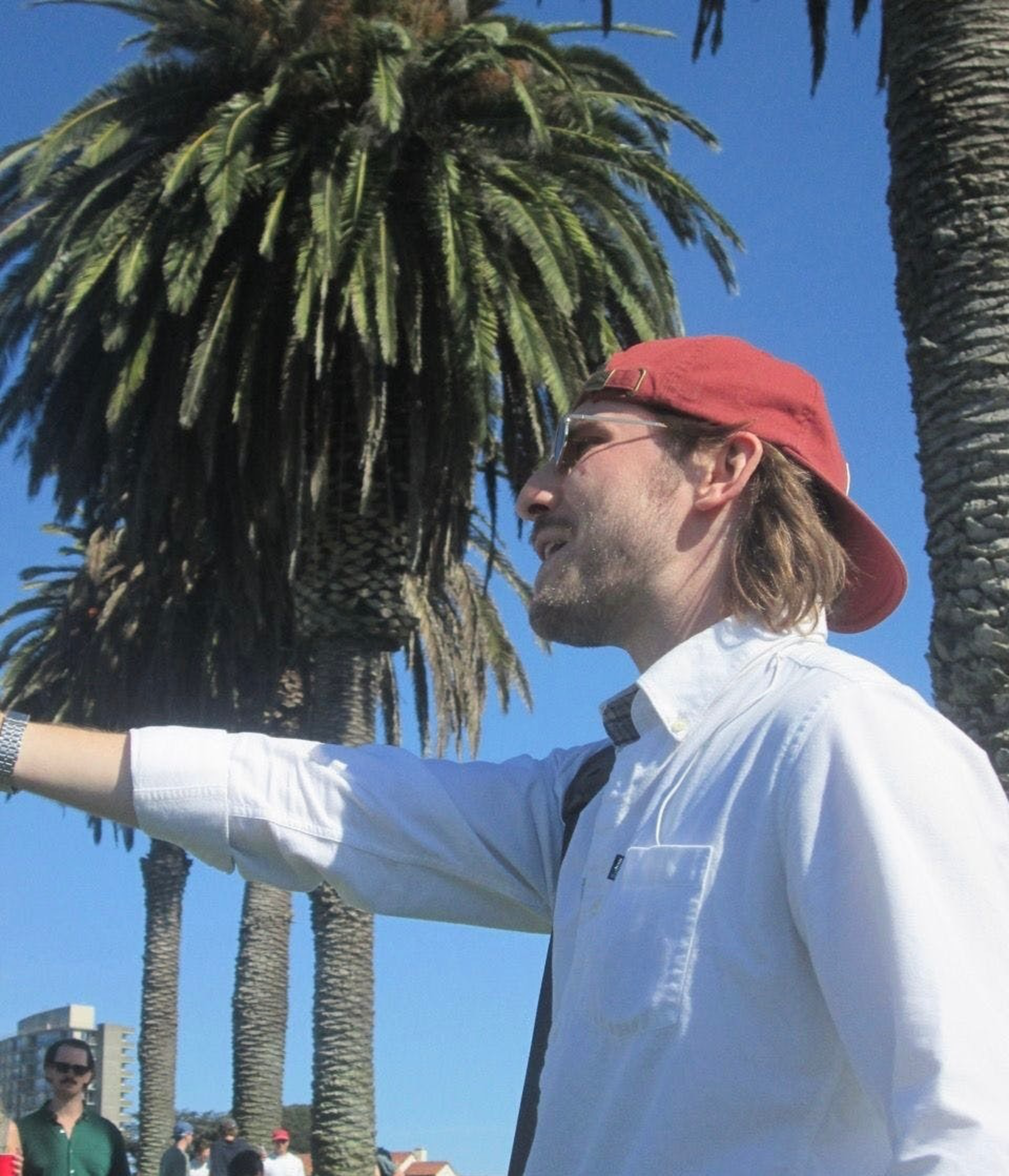 A man wearing a red backward cap, sunglasses, and white shirt reaches out with his arm, standing by tall palm trees under a clear blue sky.