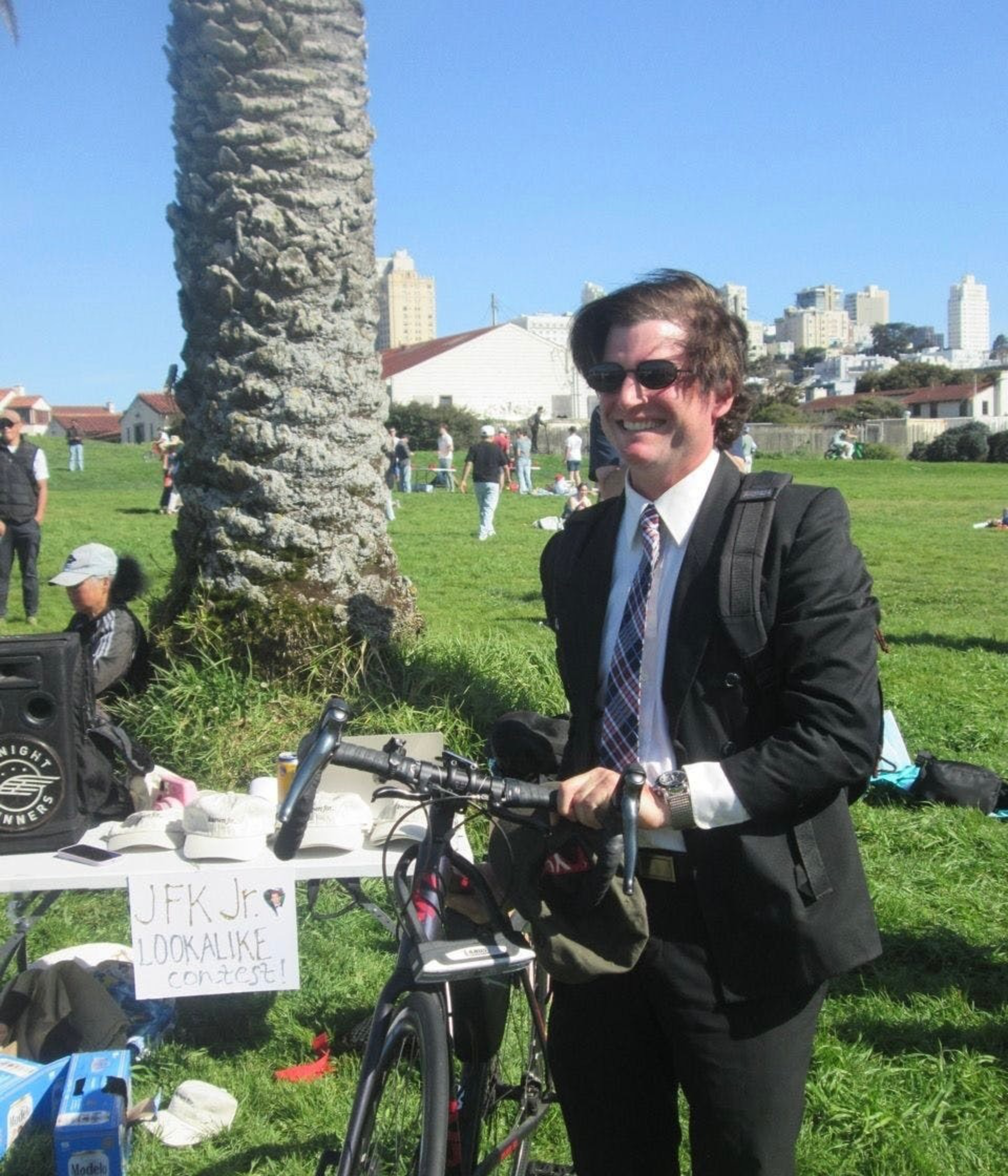 A man in a suit, tie, and sunglasses smiles while holding a bicycle at a park event labeled “JFK Jr. Lookalike Contest.”