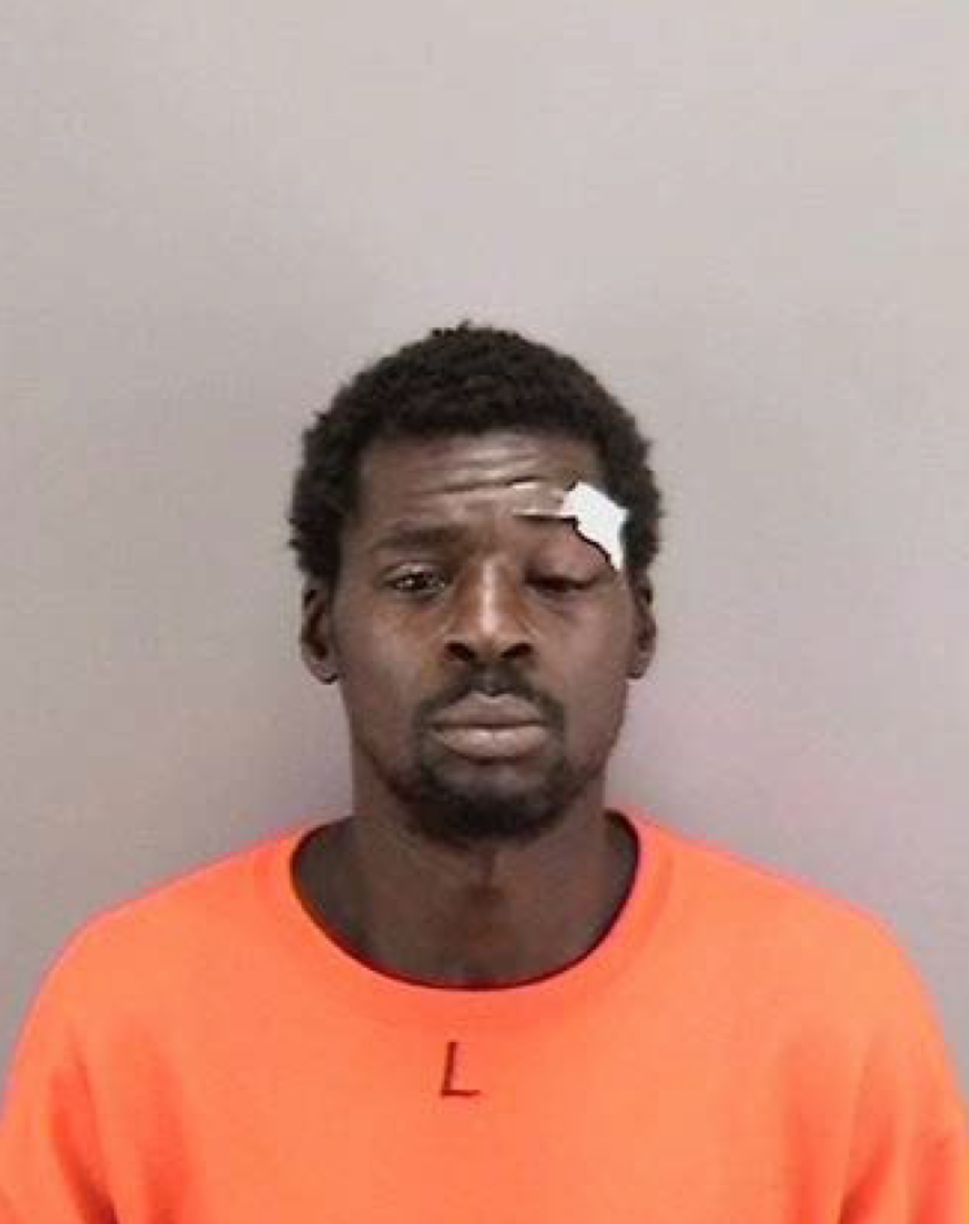 A man wearing an orange shirt with a small “L” on it has a bandage above his left eye and a neutral expression, against a plain gray background.