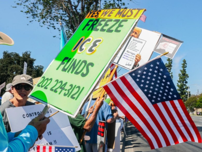 At a gathering, a man holds a sign supporting efforts to curb ICE – it reads "Freeze ICE funds" – with an American flag appearing behind it.