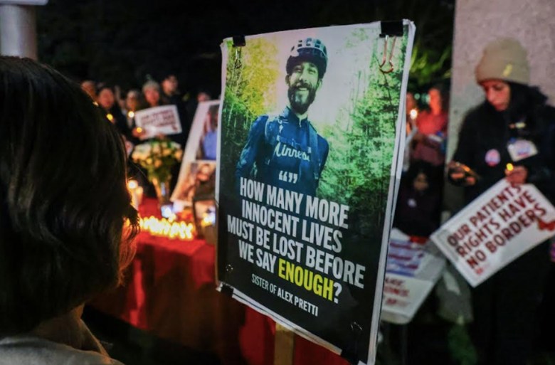 A nighttime vigil with a poster of a man and a message, surrounded by people, candles, and signs.