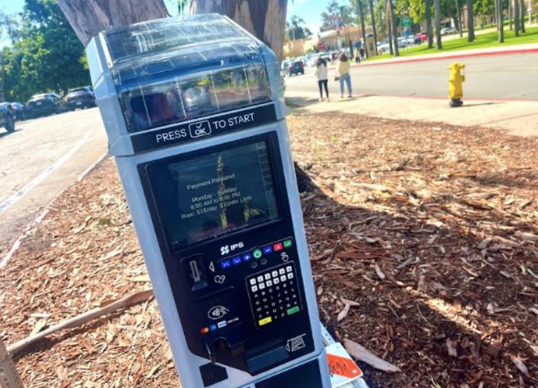 A tall machine placed in a dirt-lined patch with the bright sun glinting off it.