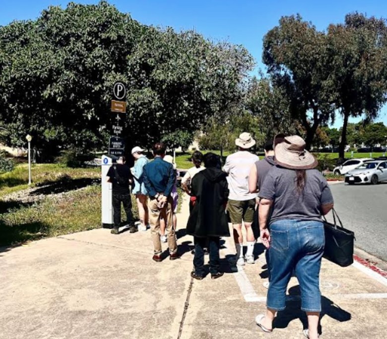 Trees form a backdrop and several people, some in hats, line up on the sidewalk for a turn at paying for parking at a kiosk. 