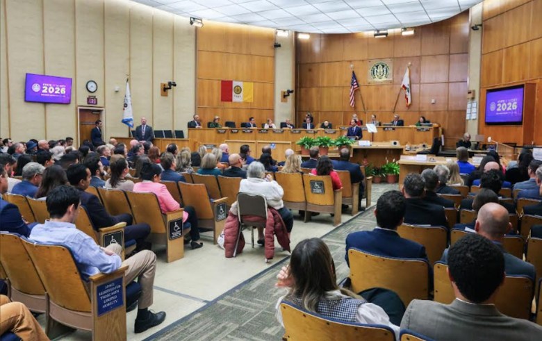 Dozens of people in wood-backed seats on either side of an aisle watch a speech by a man standing behind a dais with several other people seated on a stage behind him. Two flags are on the wall behind them, for the U.S. and California.