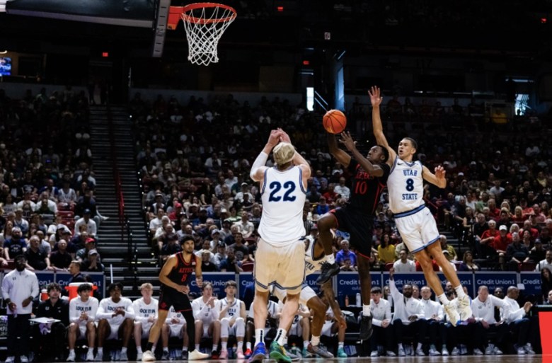 A basketball player in black takes a shot from the paint while two defenders in white jump to try and block the shot.