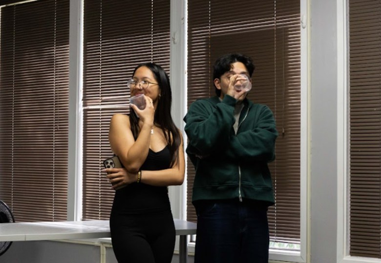 A woman in glasses and a man stand side by side on a stage rehearsing a play. The woman holds a phone and a plastic cup while the man drinks from a cup.